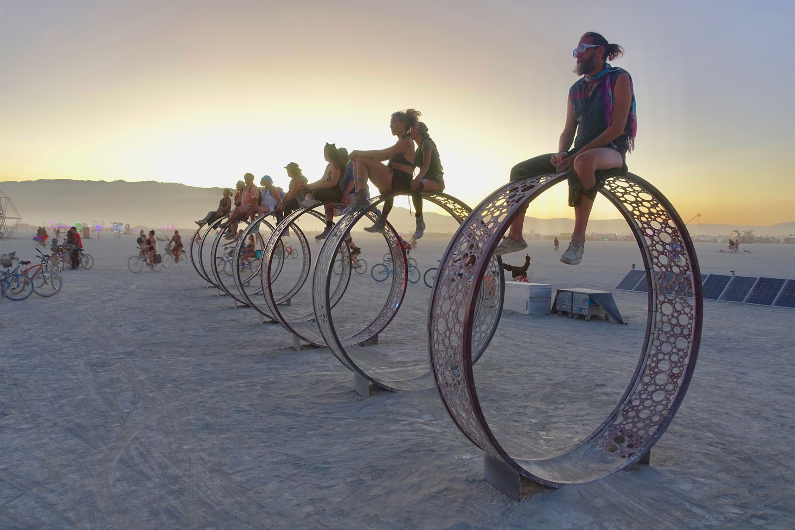Art cars and cyclists crossing the dusty open playa of Black Rock City at Burning Man, with the desert mountains beyond
